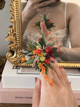 A person wears a Customized Basic Flower Ring by CAMELLIA, featuring colorful flowers and greenery, perfectly reflected in a decorative framed mirror. This adjustable piece of jewelry is expertly handcrafted in New York.